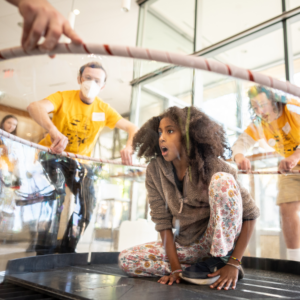Preteen crouches inside a huge soap bubble as volunteers raise the hoop around them.
